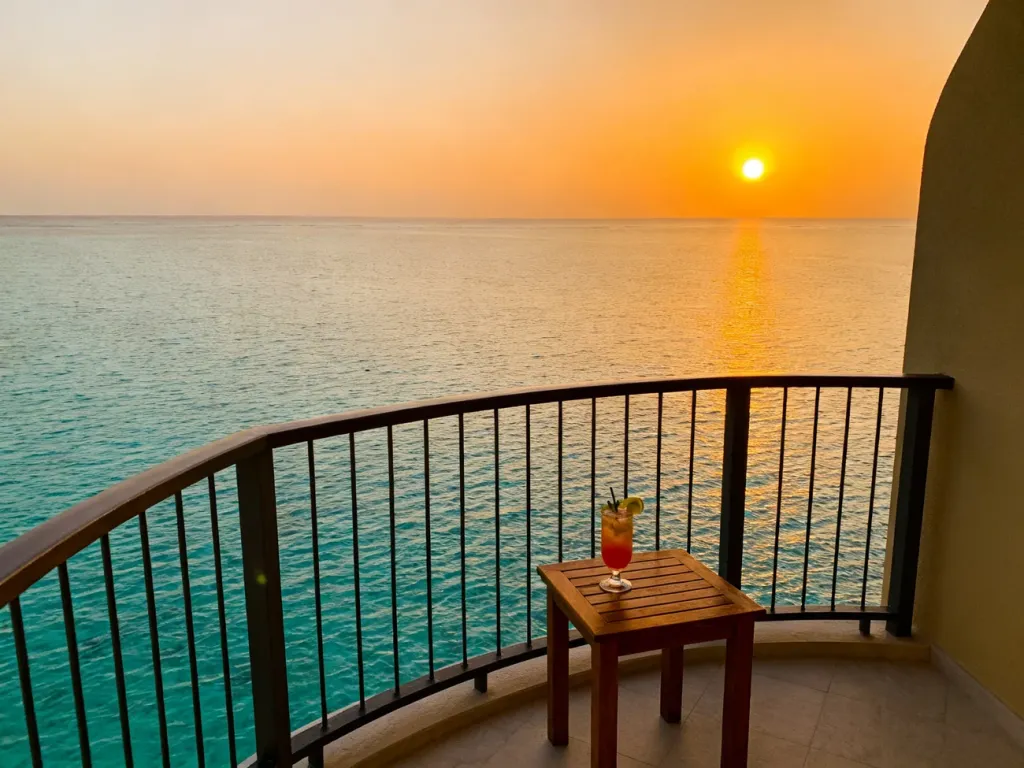 Luksusowy hotelowy balkon z widokiem na turkusowy ocean podczas zachodu słońca, ilustrujący standard pokoju typu „sea view”.